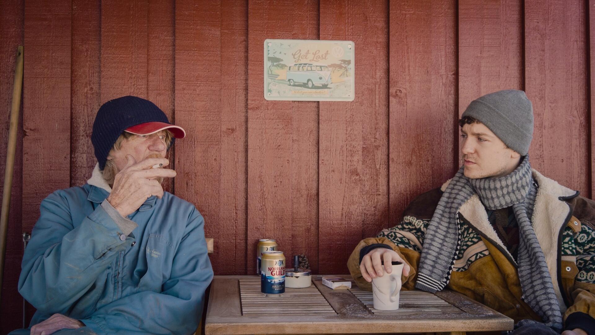 En äldre man och en yngre vid ett bord. Den äldre röker en cigarett och har en ölburk framför sig på bordet. Den yngre håller en kaffekopp. De tittar på varandra.
