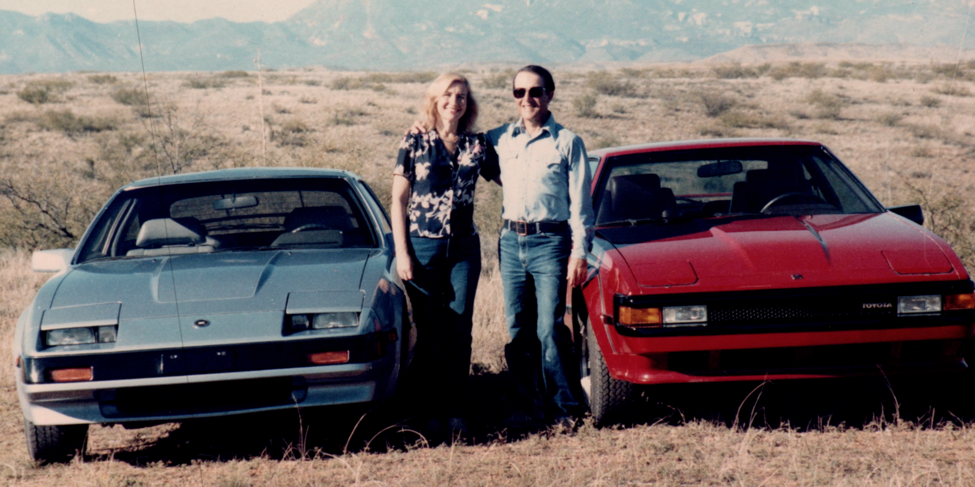 Två personer som står mellan två parkerade bilar med armarna om varandras axlar.